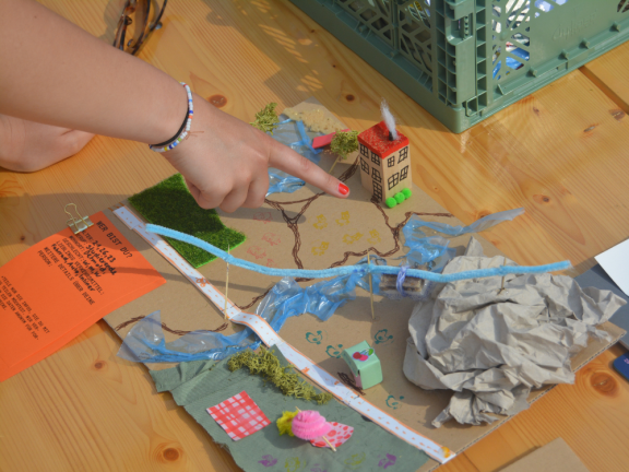 Das Foto zeigt eine rechts Frauenhand mit pinkem Nagellack, die auf einen Holztisch mit einem gebastelten Modell einer Stadt zeigt. 