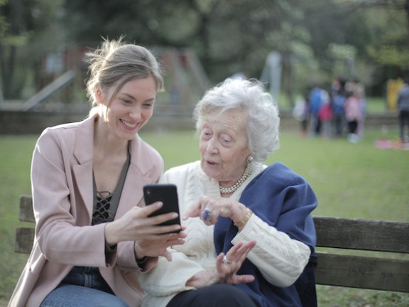 Das Symbolbild zeigt eine junge Frau auf einer Parkbank, die einer älteren Frau auf einer Parkbank etwas auf einem Smartphone zeigt. Beide lächeln. 